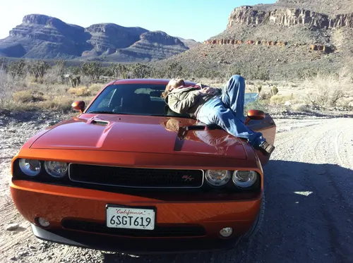 Esteban Rey descansa sobre un Dodge Challenger con su característica prenda naranja en un desierto soleado.